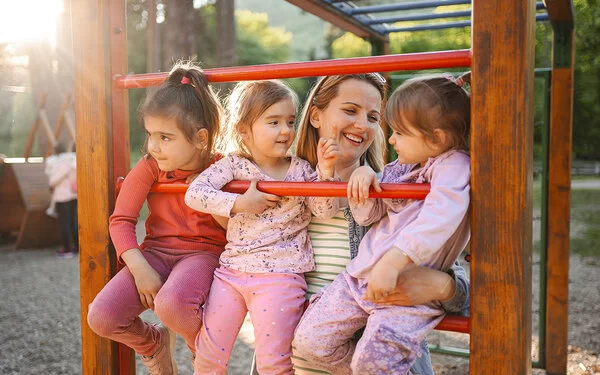 Drei Kinder sitzen lächelnd auf Klettergerüst, von einer Frau gehalten