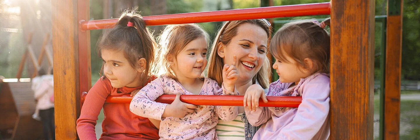 Drei Kinder sitzen lächelnd auf Klettergerüst, von einer Frau gehalten