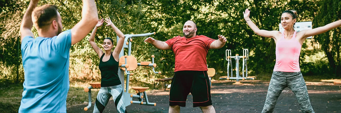 Zwei Frauen und ein Mann sind draußen in einem Bewegungspark und machen Hampelmänner. Ein Trainer, der nur von hinten zu sehen ist, zeigt ihnen, wie es geht.