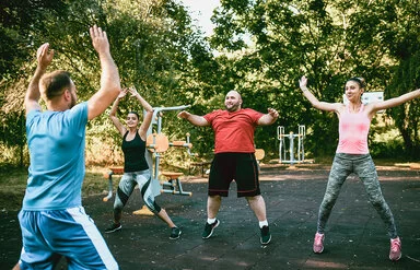 Zwei Frauen und ein Mann sind draußen in einem Bewegungspark und machen Hampelmänner. Ein Trainer, der nur von hinten zu sehen ist, zeigt ihnen, wie es geht.