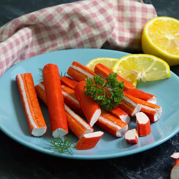 Surimi-Stäbchen liegen auf einem blauen Teller, garniert mit Zitronenscheiben und etwas Petersilie.