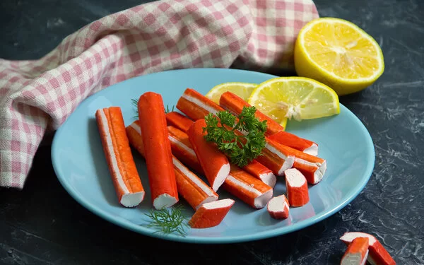 Surimi-Stäbchen liegen auf einem blauen Teller, garniert mit Zitronenscheiben und etwas Petersilie.