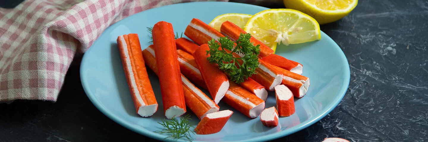 Surimi-Stäbchen liegen auf einem blauen Teller, garniert mit Zitronenscheiben und etwas Petersilie.