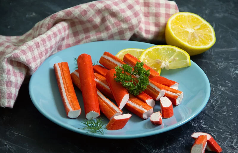 Surimi-Stäbchen liegen auf einem blauen Teller, garniert mit Zitronenscheiben und etwas Petersilie.