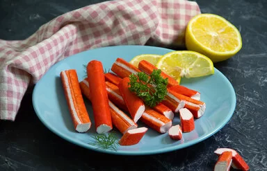 Surimi-Stäbchen liegen auf einem blauen Teller, garniert mit Zitronenscheiben und etwas Petersilie.