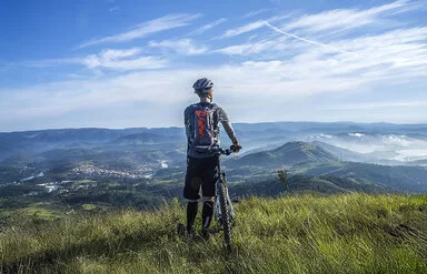 Rückenansicht eines Radfahrers mit einem Mountainbike. Er steht auf einer sommerlichen Wiese und blickt auf das vor ihm liegende Tal und eine Bergkette. Der Radfahrer trägt hohe schwarze Socken, eine schwarze Sporthose, ein graues langärmeliges Funktionsshirt, einen blau-roten Rucksack und einen grau-blauen Helm.