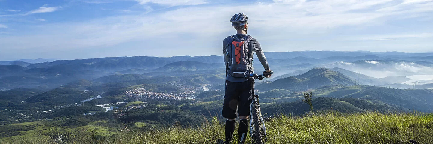 Rückenansicht eines Radfahrers mit einem Mountainbike. Er steht auf einer sommerlichen Wiese und blickt auf das vor ihm liegende Tal und eine Bergkette. Der Radfahrer trägt hohe schwarze Socken, eine schwarze Sporthose, ein graues langärmeliges Funktionsshirt, einen blau-roten Rucksack und einen grau-blauen Helm.