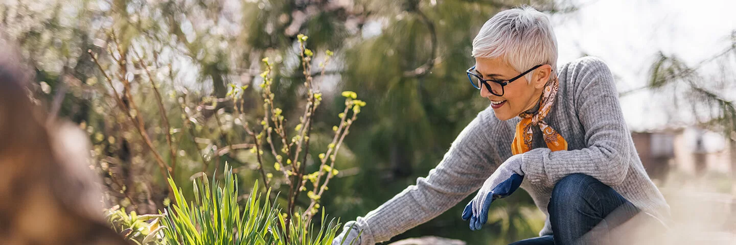 Eine ältere Frau mit einem grauen Pullover und einem Halstuch kniet neben einem Beet und arbeitet mit Gartenwerkzeugen.