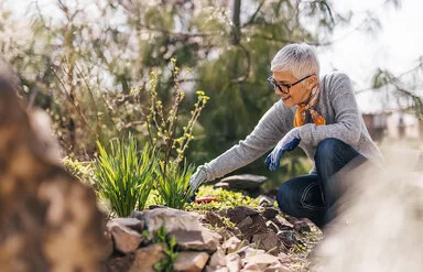 Eine ältere Frau mit einem grauen Pullover und einem Halstuch kniet neben einem Beet und arbeitet mit Gartenwerkzeugen.