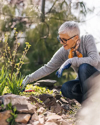 Eine ältere Frau mit einem grauen Pullover und einem Halstuch kniet neben einem Beet und arbeitet mit Gartenwerkzeugen.