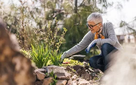 Eine ältere Frau mit einem grauen Pullover und einem Halstuch kniet neben einem Beet und arbeitet mit Gartenwerkzeugen.