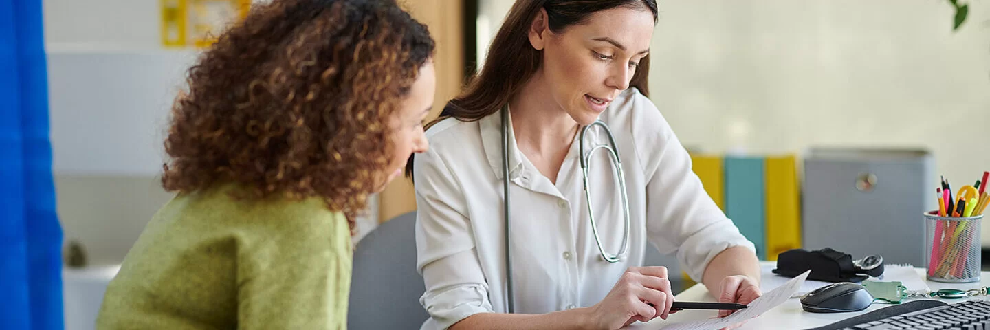 Eine Ärztin sitzt mit einer Patientin an einem Schreibtisch und zeigt mit einem Kugelschreiber auf ein Dokument. 