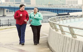 Zwei übergewichtige Personen walken und unterhalten sich dabei. Im Hintergrund sind ein Fluss und eine Brücke sowie Bürogebäude zu sehen.