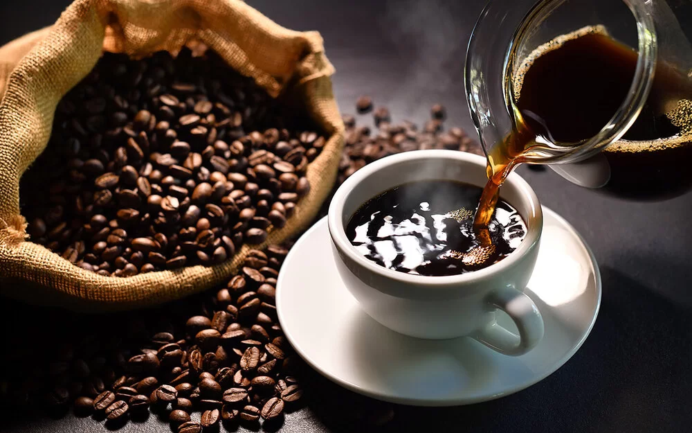 Schwarzer Kaffee wird aus einem Glaskrug in eine weiße Tasse mit Untersetzer gegossen. Daneben liegt ein Sack mit Kaffeebohnen.