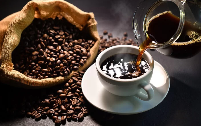 Schwarzer Kaffee wird aus einem Glaskrug in eine weiße Tasse mit Untersetzer gegossen. Daneben liegt ein Sack mit Kaffeebohnen.