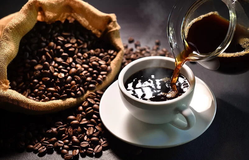 Schwarzer Kaffee wird aus einem Glaskrug in eine weiße Tasse mit Untersetzer gegossen. Daneben liegt ein Sack mit Kaffeebohnen.