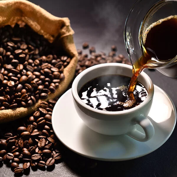 Schwarzer Kaffee wird aus einem Glaskrug in eine weiße Tasse mit Untersetzer gegossen. Daneben liegt ein Sack mit Kaffeebohnen.