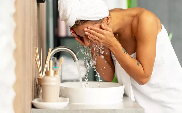 Badezimmer. Eine Frau hat sich ein weißes Handtuch um den Körper geschlungen. Um die Haare hat sie ein weißes Handtuch als Turban gewickelt. Sie spritzt sich am Waschbecken bei laufendem Wasserhahn Wasser ins Gesicht.