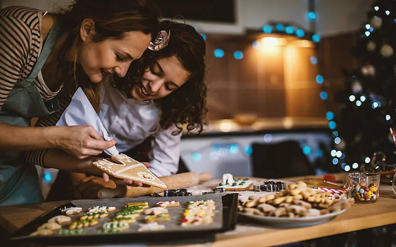 Mutter und Tochter backen zusammen für Weihnachten. Auf einem Backblech liegen Plätzchen, die mit bunten Zuckerperlen und Streuseln dekoriert sind, auf einem weißten Teller liegen Butterplätzchen. Die Mutter dekoriert mit einem Spritzbeutel ein Lebkuchenhaus. Ihre Tochter schaut freudestrahlend zu, wie sie den runden Hauseingang mit weißem Zuckerguss umrandet.