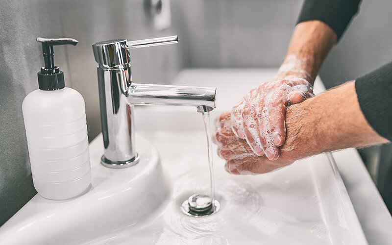 Zwei eingeseifte Hände eines älteren Mannes in einem Waschbecken mit laufendem Wasser.