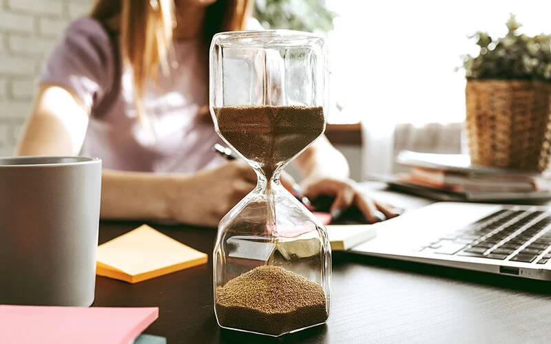 Eine Frau sitzt an einem Schreibtisch und notiert etwas auf einem Zettel. Vor ihr steht eine Sanduhr.