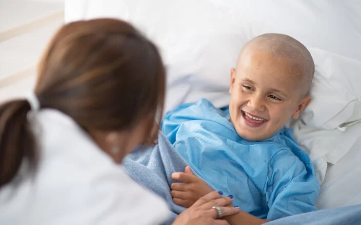 Ein kleiner Krebspatient, dem die Haare ausgefallen sind, liegt in einem Krankenhausbett. Eine Ärztin beugt sich zu ihm vor. Ihre rechte Hand liegt auf seinem linken Handgelenk. Sie bringt den Jungen zum Lachen.