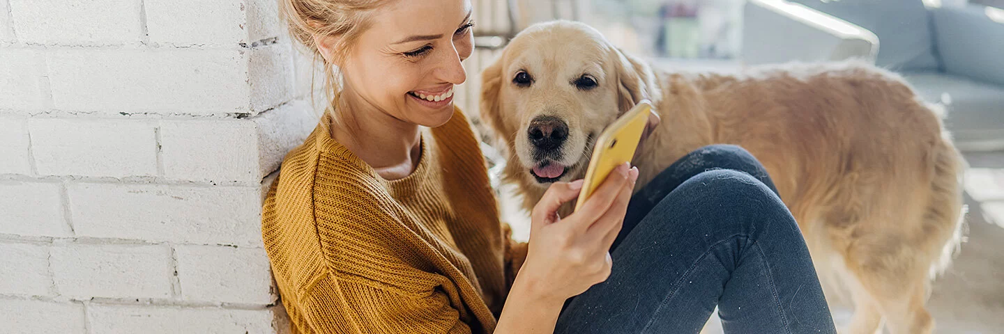 Junge Frau hockt in der Morgensonne an eine Mauer gelehnt auf dem Boden und liest etwas auf ihrem Smartphone, ein Hund steht an ihrer Seite und schaut ihr zu.