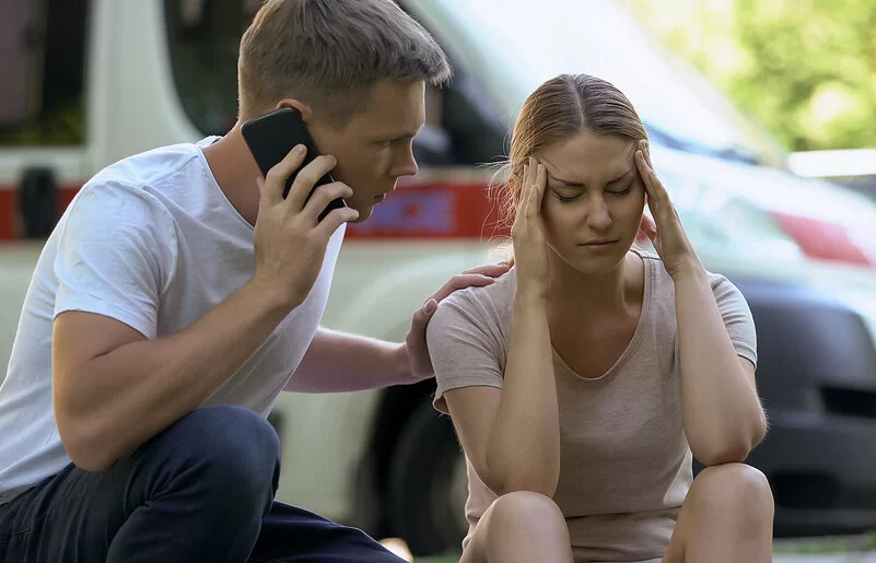 Eine junge Frau fasst sich vor Schmerzen an die Schläfen. Ihre Augen sind geschlossen. Neben ihr hockt ein junger Mann, der seine linke Hand auf ihre Schulter gelegt hat. Mit der rechten Hand hält er ein Handy an sein rechtes Ohr, um Hilfe zu rufen. Im Hintergrund sind verschwommen parkende Autos zu sehen.