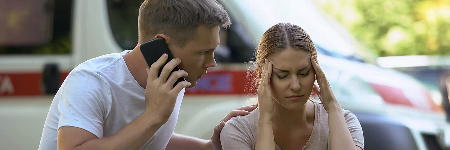 Eine junge Frau fasst sich vor Schmerzen an die Schläfen. Ihre Augen sind geschlossen. Neben ihr hockt ein junger Mann, der seine linke Hand auf ihre Schulter gelegt hat. Mit der rechten Hand hält er ein Handy an sein rechtes Ohr, um Hilfe zu rufen. Im Hintergrund sind verschwommen parkende Autos zu sehen.