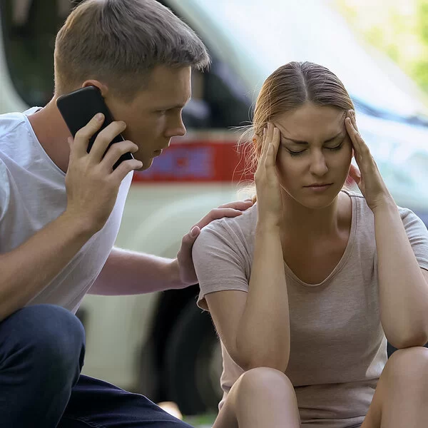 Eine junge Frau fasst sich vor Schmerzen an die Schläfen. Ihre Augen sind geschlossen. Neben ihr hockt ein junger Mann, der seine linke Hand auf ihre Schulter gelegt hat. Mit der rechten Hand hält er ein Handy an sein rechtes Ohr, um Hilfe zu rufen. Im Hintergrund sind verschwommen parkende Autos zu sehen.