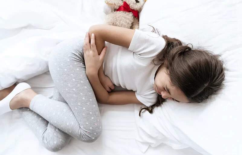 Ein Mädchen liegt im Bett und hält sich mit schmerzverzerrtem Gesicht die Arme vor den Bauch.