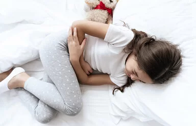 Ein Mädchen liegt im Bett und hält sich mit schmerzverzerrtem Gesicht die Arme vor den Bauch.