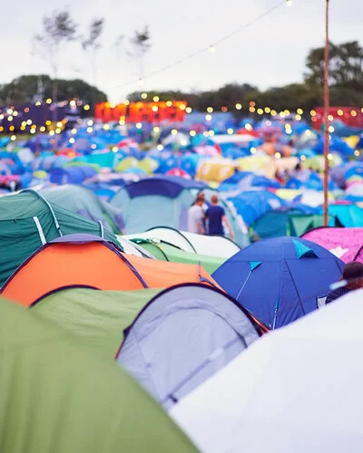 Aufnahme eines Campingplatzes mit vielen bunten Zeltdächern auf einem Outdoor-Festival. Über den Zelten hängen Lichterketten.