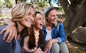 Ein Mädchen sitzt zwischen zwei älteren Frauen, alle lachen gemeinsam.