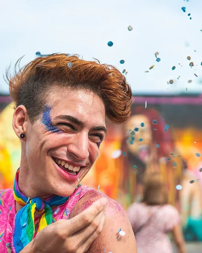 Porträt eines lachenden queeren Mannes auf einem Festival, der blauen Glitzer neben seinem rechten Auge trägt und Konfetti in die Luft wirft.