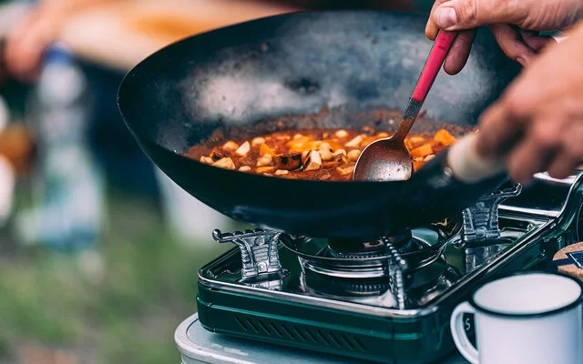 Eine nicht sichtbare Person rührt mit einem Löffel in einem Eintopf in einer Wokpfanne auf einem Campingkocher.