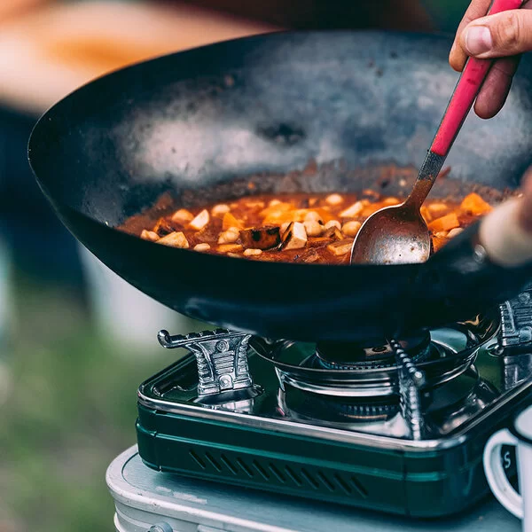 Eine nicht sichtbare Person rührt mit einem Löffel in einem Eintopf in einer Wokpfanne auf einem Campingkocher.