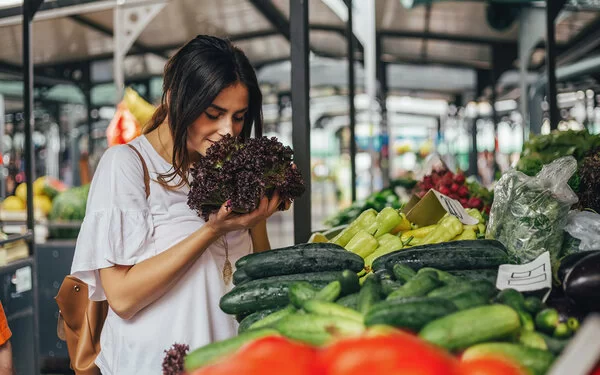 Junge Frau riecht an einem Salat auf einem Markt mit viel Gemüse. Bildunterschrift: Frisches Gemüse vom Markt ist Balsam für die Seele.