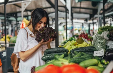 Junge Frau riecht an einem Salat auf einem Markt mit viel Gemüse. Bildunterschrift: Frisches Gemüse vom Markt ist Balsam für die Seele.