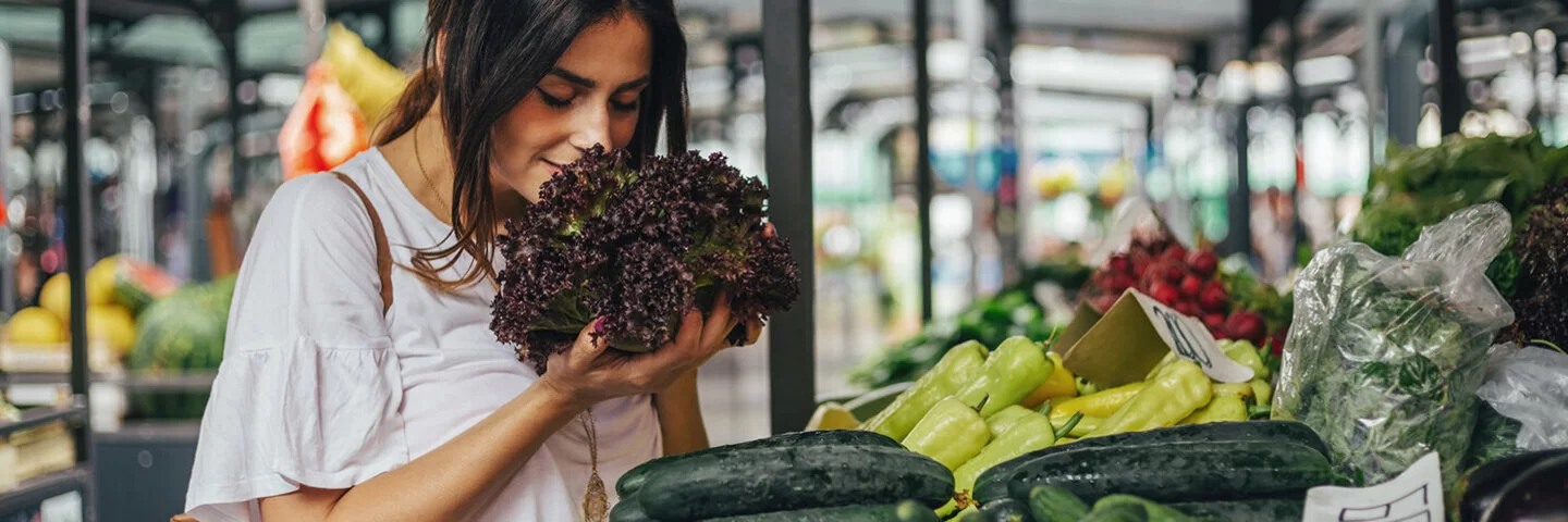 Junge Frau riecht an einem Salat auf einem Markt mit viel Gemüse. Bildunterschrift: Frisches Gemüse vom Markt ist Balsam für die Seele.