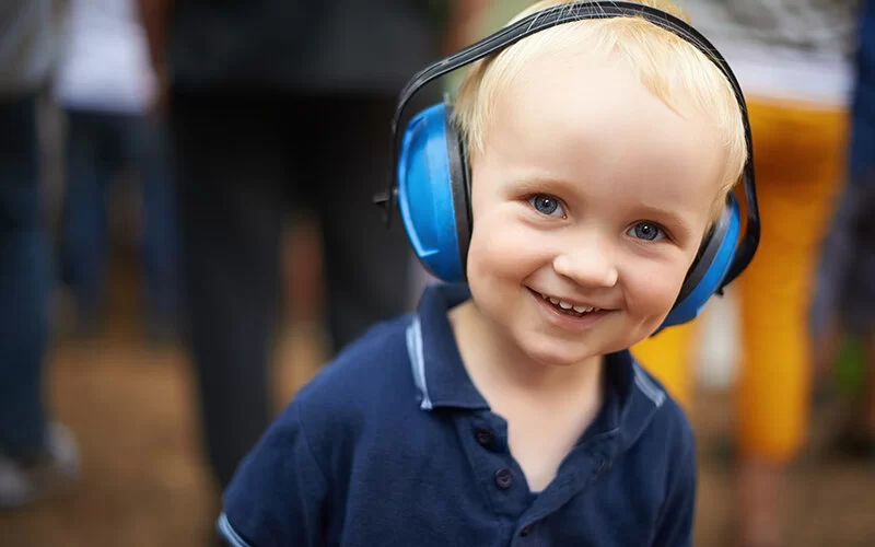 Ein lächelnder kleiner Junge mit blonden Haaren, der ein dunkelblaues Poloshirt trägt und blaue Ohrschützer aufhat.