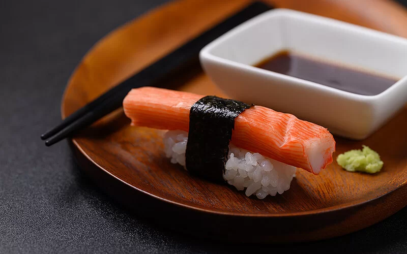 Ein Nigiri-Sushi mit Surimi liegt auf einem Holzteller. Neben dem Sushi sieht man eine Schale Sojasauce, schwarze Essstäbchen und ein Klecks Wasabi.