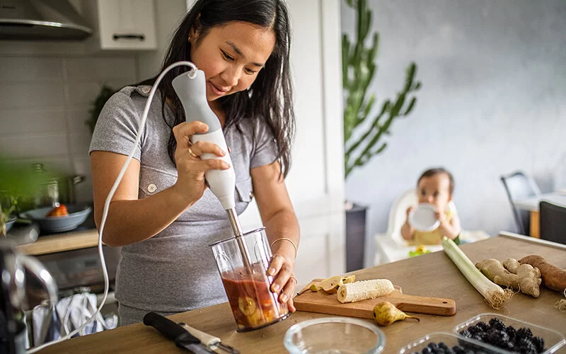 Eine Frau steht in einer Küche an einem Tisch und püriert mit einem Pürierstab Bananen, Birnen und Heidelbeeren. Auf dem Tisch liegt außerdem frisches Gemüse. Im Hintergrund ist ihr Baby zu sehen.