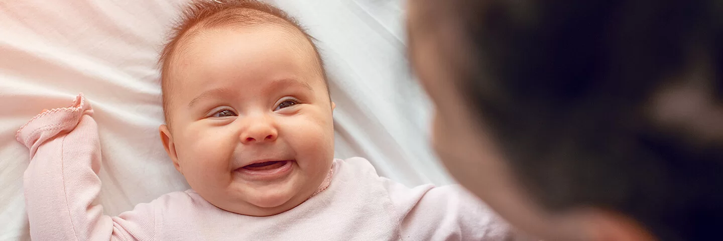 Ein kleines Baby in einem rosa Strampler liegt auf einem weißen Laken und lächelt.  Am oberen Bildrand ist verschwommen der Hinterkopf einer dunkelhaarigen Frau zu erkennen.