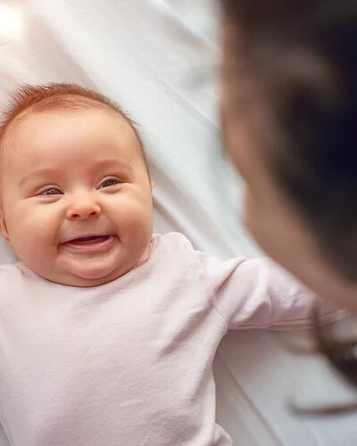 Ein kleines Baby in einem rosa Strampler liegt auf einem weißen Laken und lächelt.  Am oberen Bildrand ist verschwommen der Hinterkopf einer dunkelhaarigen Frau zu erkennen.