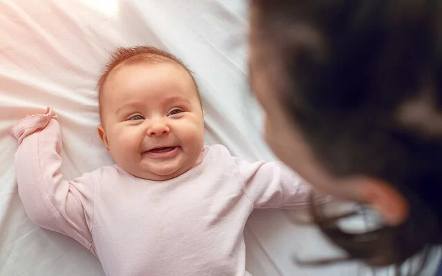 Ein kleines Baby in einem rosa Strampler liegt auf einem weißen Laken und lächelt.  Am oberen Bildrand ist verschwommen der Hinterkopf einer dunkelhaarigen Frau zu erkennen.