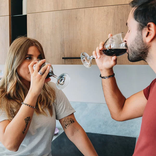 Eine junge Frau und ein junger Mann trinken Rotwein in der Küche. Sie trägt ihre langen, dunkelblonden Haare in der Mittte gescheitelt und offen. An ihren beiden Armen ist jeweils ein schwarzes Tattoo zu sehen. Er hat kurze schwarze Haare, einen kurzen Vollbart und trägt ein rotes T-Shirt. Die beiden schauen einander an.