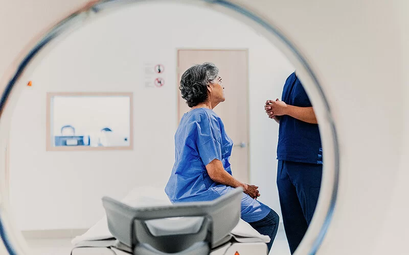 Eine ältere Patientin spricht mit einer medizinischen Fachkraft vor einem Magnetresonanztomographen. Das Bild wurde aus der Röhre des MRT-Gerätes aufgenommen, deren Umrisse deutlich erkennbar sind.