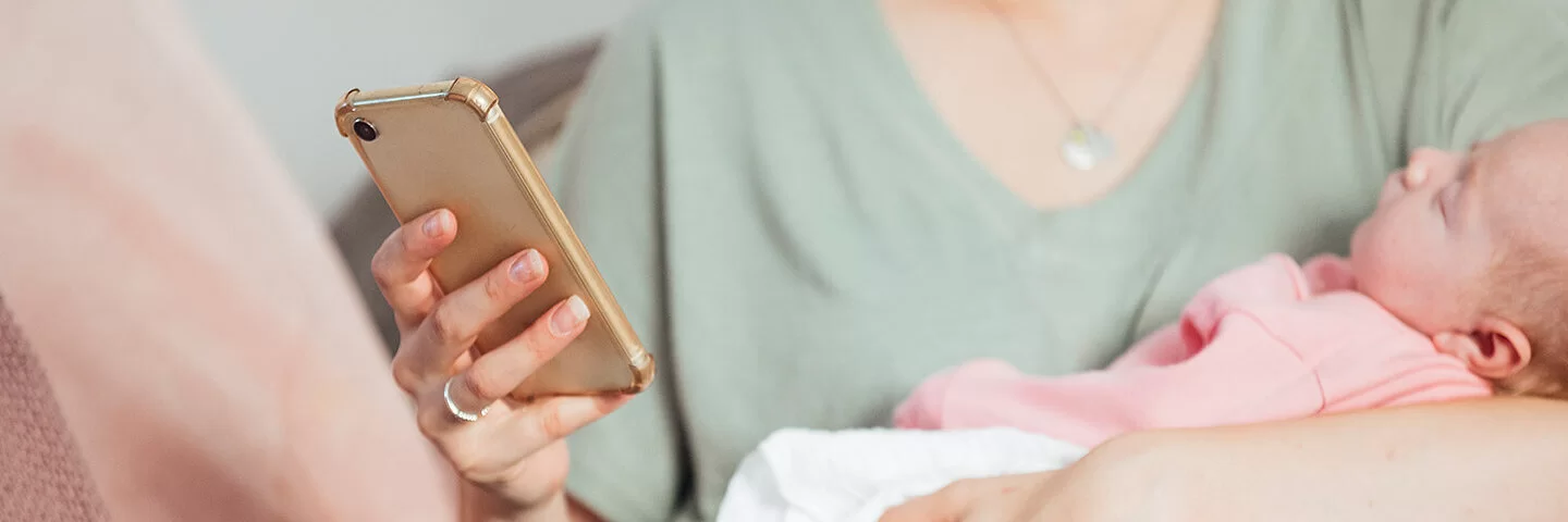 Eine Mama im hellgrünen T-Shirt hält ihren schlafenden Säugling im Arm. In der anderen Hand hält die Frau ihr Smartphone und schaut etwas darauf nach.