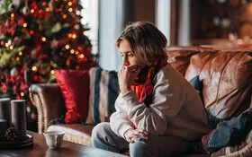 Eine Jugendliche sitzt traurig auf einem Sofa im Wohnzimmer. Sie stützt ihr Kinn auf die linke Hand. Im Hintergrund steht ein erleuchteter Weihnachtsbaum.